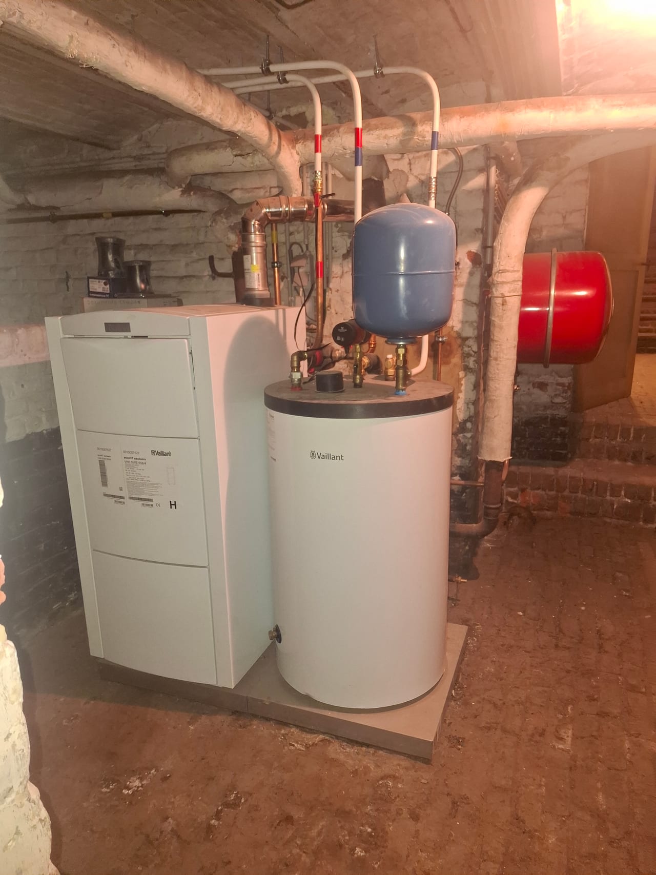 Chaudière au sol Vaillant et ballon d'eau chaude, vases d'expansion bleu et rouge et conduites dans une cave en briques.