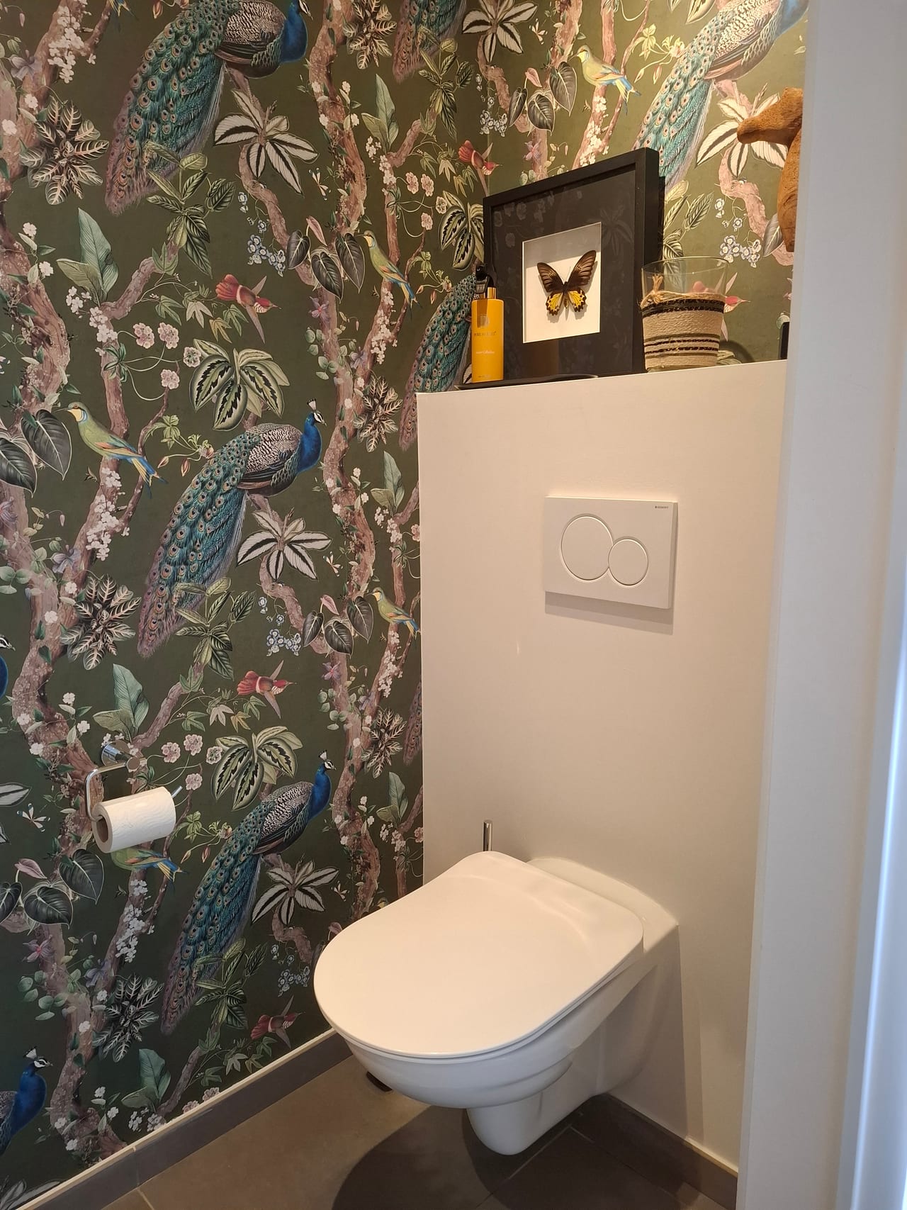 WC suspendu blanc avec commande Geberit à double touche, devant un papier peint à motifs de paons.