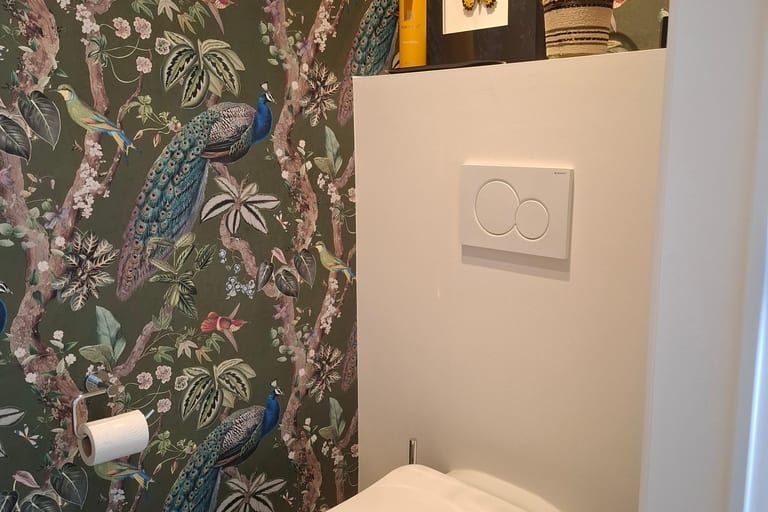 Plaque de commande Geberit à double touche sur un WC suspendu réparé par Atmos Technics à Bruxelles, mur tapissé d'un papier peint à motifs de paons.